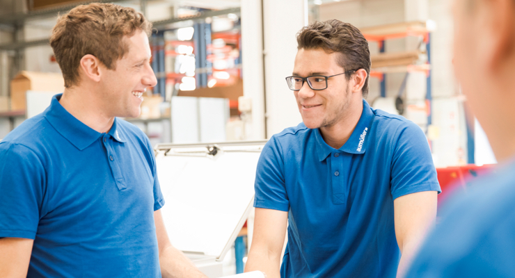 Das Bild zeigt zwei Männer in blauer Unternehmensbekleidung die sich anlächeln