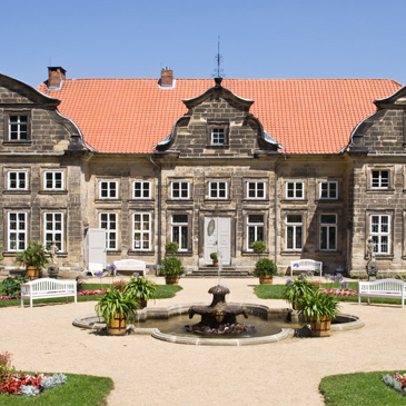Auf dem Bild ist der Eingang des Schloss Blankenburg zu sehen vor dem grauen Haus mit rotem Schrägdach ist ein Platz um einem Brunnen