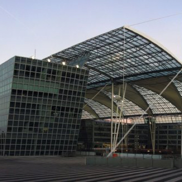 Auf dem Bild  ist das Terminal 2 des Flughafen München zu sehen. Ein Glasgebäude unter einem geschwungenen Glasdach zu sehen